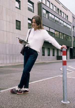 Vrouw in witte trui en spijkerbroek leest een krant, leunend tegen een rood-wit gestreepte straatpaal op een stedelijk trottoir.