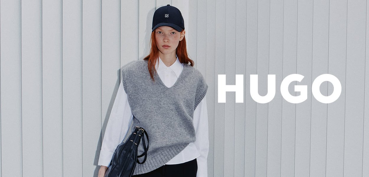 Szary, dziergany, bezrękawnik noszony na białej koszuli z kołnierzykiem, zestawiony z granatową czapką bejsbolową, trzymający czarną torebkę. Pionowe, paskowane tło.