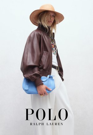 Vrouw met brede hoed, bruine leren jas, witte broek, die een lichtblauwe handtas vasthoudt tegen een effen witte achtergrond.