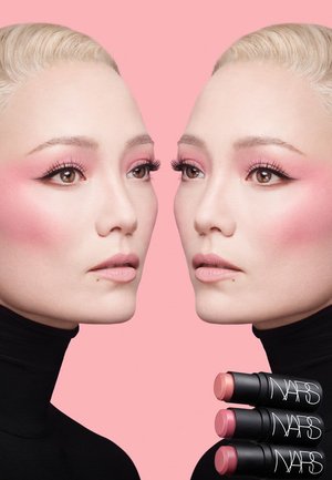 Due modelle con capelli biondo chiaro e trucco rosa leggero che si guardano l'una con l'altra, su uno sfondo rosa. Tre tubetti di rossetto NARS in primo piano.