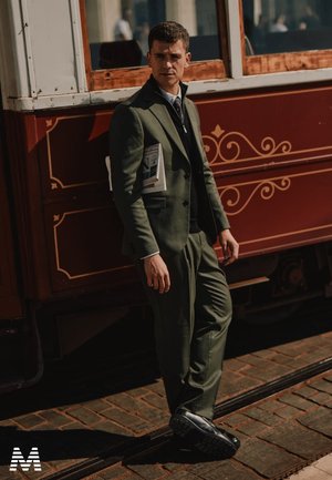 Dunkelgrüner Anzug mit taillierter Passform und Revers, über einer schwarzen Weste getragen. Schwarze Schuhe mit strukturierten Sohlen. Hält eine Zeitschrift und posiert vor einer historischen Straßenbahn.