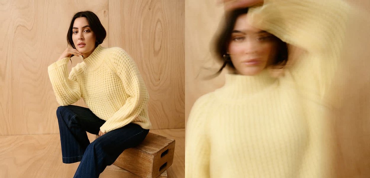 Frau mit dunklen Haaren, die einen gelben Strickpullover und dunkle Jeans trägt, sitzt auf einer Holzkiste vor einer hellen Holzwand.