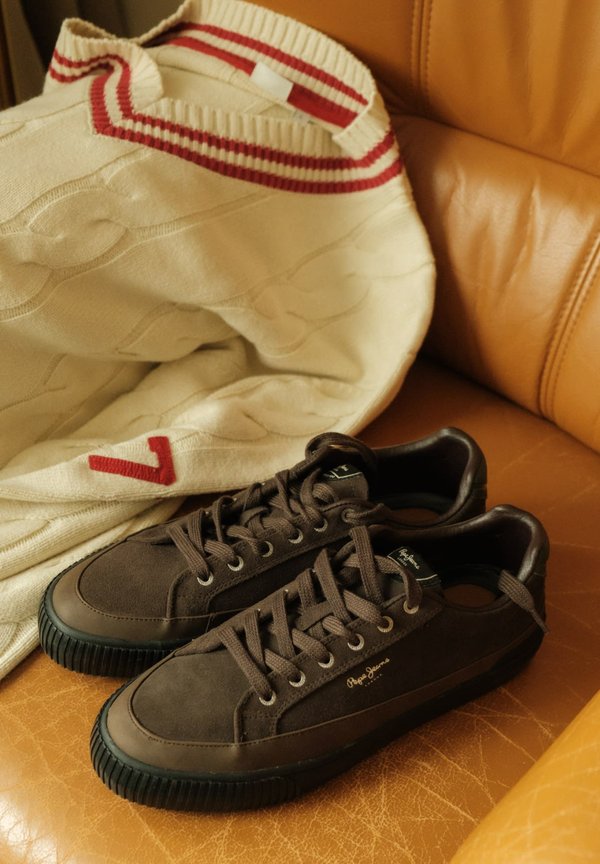 Zapatillas de ante marrón con suelas de goma negra, cordones y ojetes plateados. En el fondo hay un suéter de punto crema con rayas rojas y un patrón de trenzado.