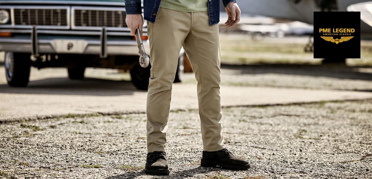 Beige stretch katoenen broek met een slanke pasvorm, gecombineerd met zwarte schoenen en een groene shirt, met een moersleutel in een robuuste buitenomgeving.