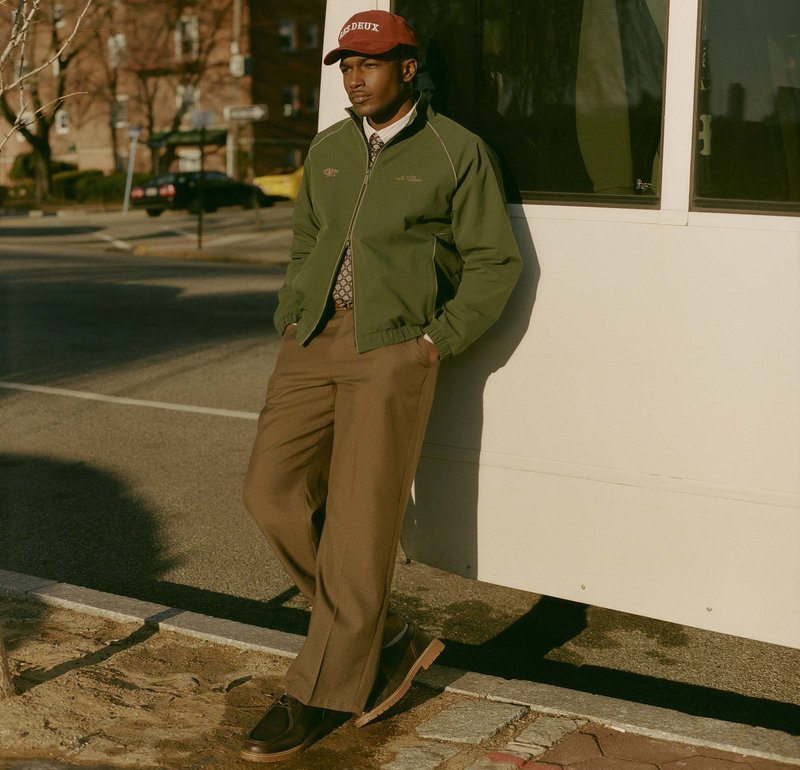 Grøn bomberjakke, mønstret slips, brune bukser og mørke sko. Mand læner sig op ad en hvid væg, skygge synlig på jorden.