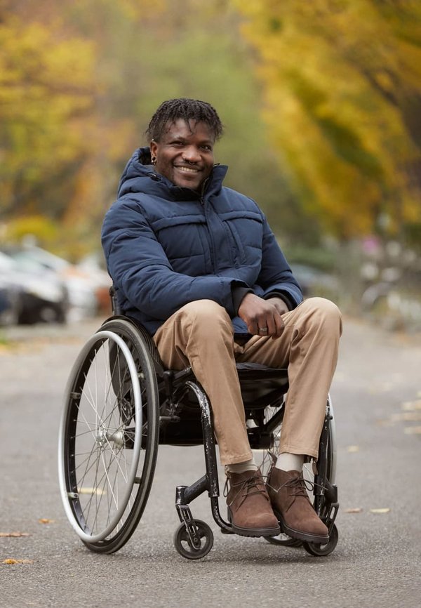 Manuell rullstol med stora hjul, svart säte och silverdetaljer. Person klädd i en blå vadderad jacka, beige byxor och bruna skor.