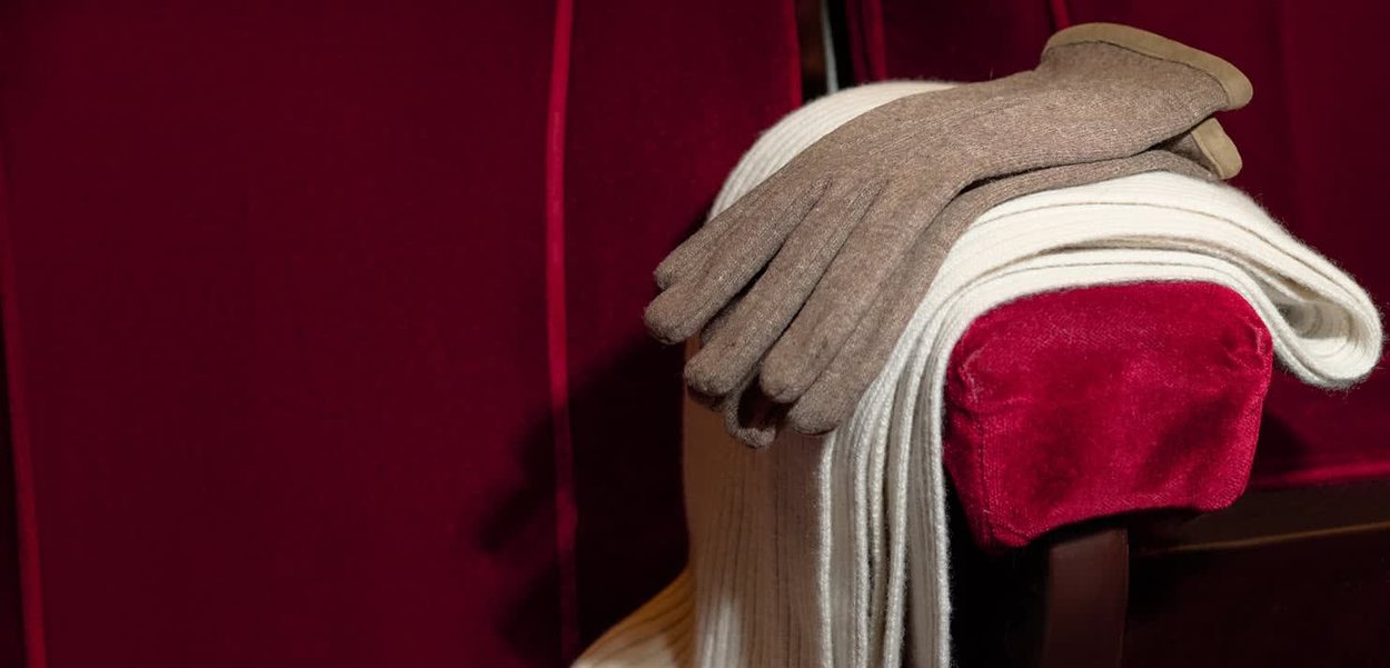Guanti di lana marrone riposano su una sciarpa color crema piegata, posta su una sedia rivestita di tessuto rosso scuro. Le texture sono morbide e calde.