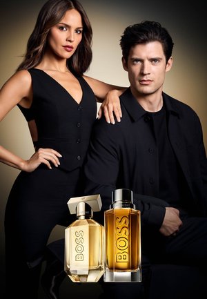 Donna e uomo vestiti di nero posano dietro due bottiglie di profumo BOSS color ambra etichettate "The Scent".