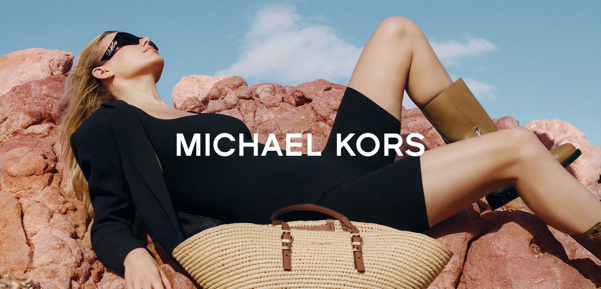 Black bodycon dress, oversized black blazer, knee-length brown boots, straw handbag with a brown strap, resting on rocky terrain.