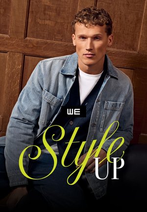 Junger Mann mit lockigem Haar, der eine Jeansjacke, ein weißes Hemd und eine dunkle Weste trägt, sitzt vor einer holzvertäfelten Wand.