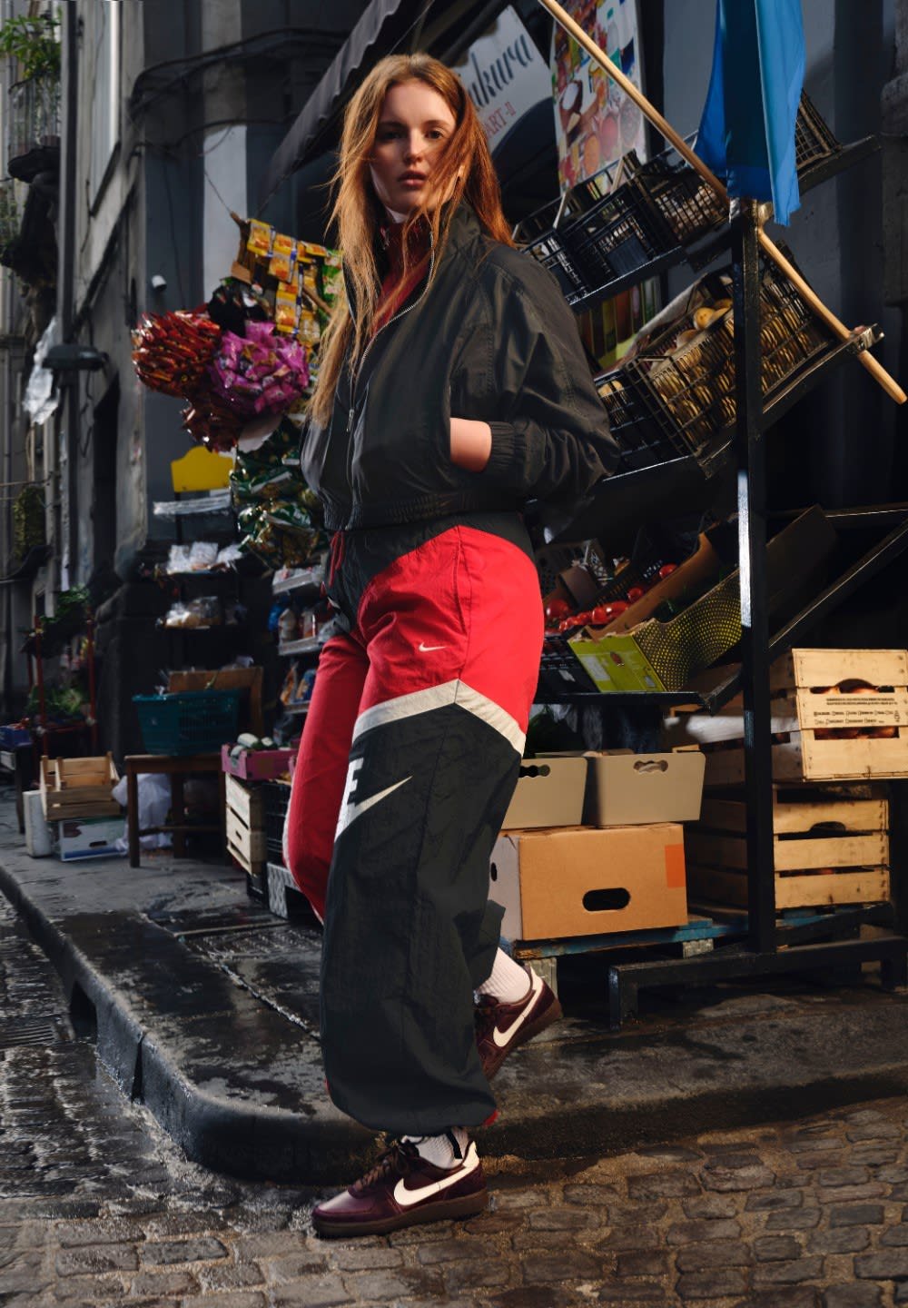 Nera giacca bomber, pantaloni sportivi rossi e neri con accenti bianchi, indossati con sneakers scure. Ambientazione urbana con sfondo di mercato.