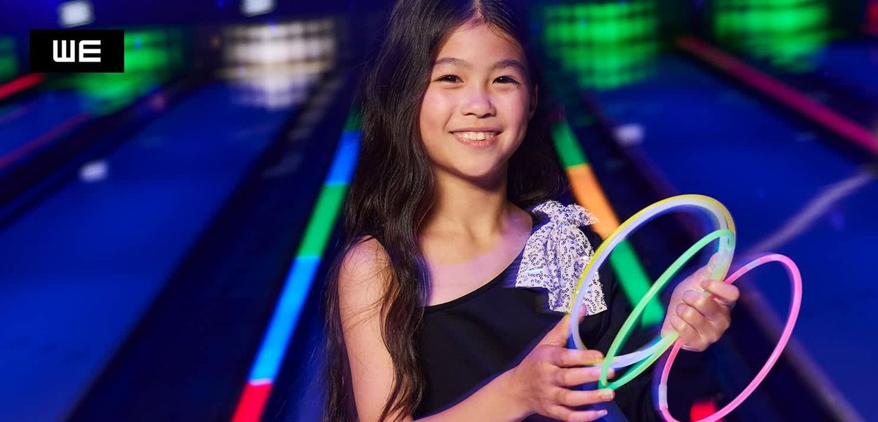 Ein Mädchen hält drei leuchtende Ringe in Neonpink, Grün und Gelb. Sie trägt ein schwarzes Oberteil mit einem silbernen, gemusterten Akzent. Bowlingbahnen sind sichtbar.