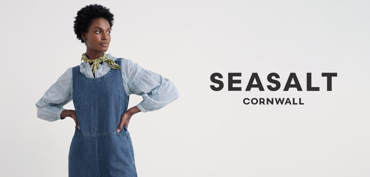 Denim overall dress with a fitted bodice and wide straps, paired with a light blue, puff-sleeve shirt and a patterned scarf.