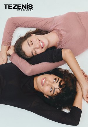 Zwei Frauen liegen auf einem hellen Hintergrund, eine in einem engen dunklen Oberteil und die andere in einem hellrosa Oberteil mit Spitzenverzierungen am Ausschnitt.