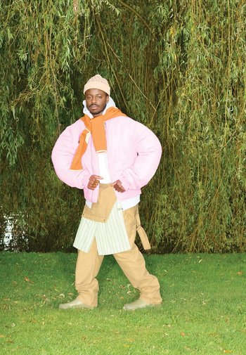 Roze pufferjack over oranje sweater, witte shirt, kaki broek en een schort met groene strepen. Staande op gras met bomen op de achtergrond.