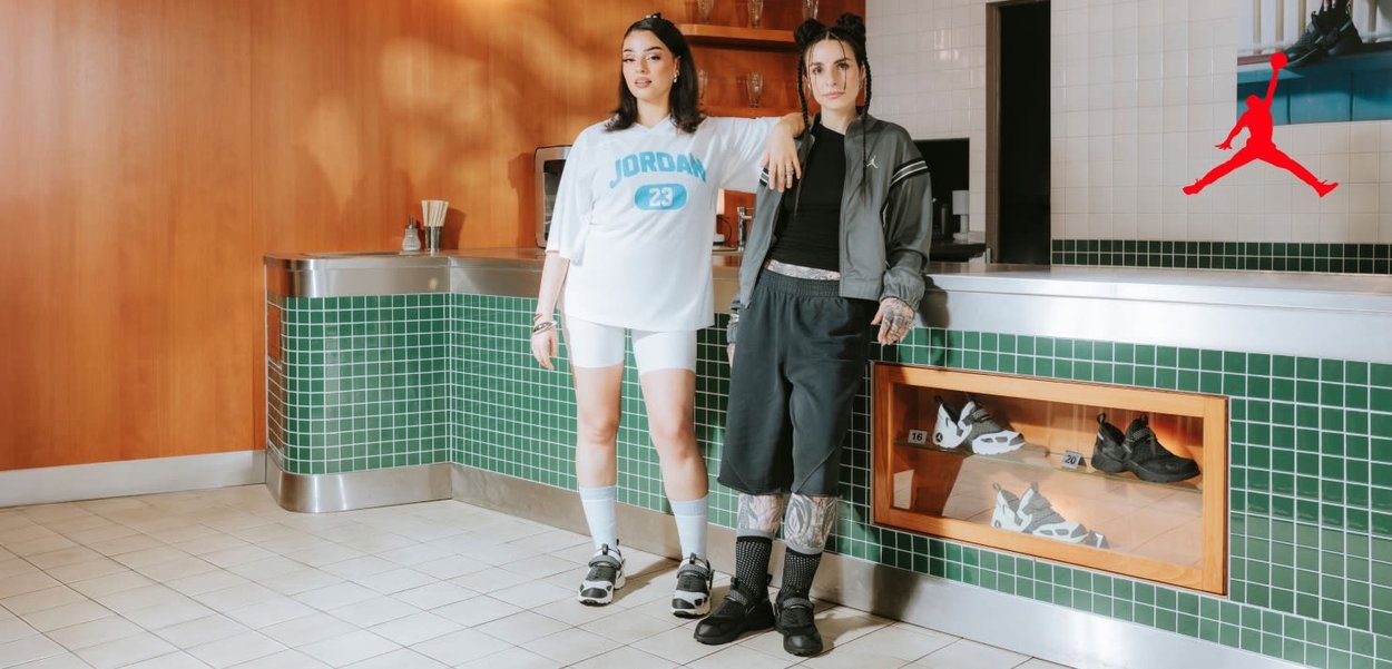 Two women stand by a green-tiled counter. One wears a white jersey with blue lettering and bike shorts; the other, a dark jacket and black shorts.