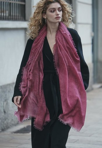 Lang, pink scarf lavet af letvægtsstof med frynset kanter, båret over et sort outfit. Modellen har krøllet hår og går udendørs.