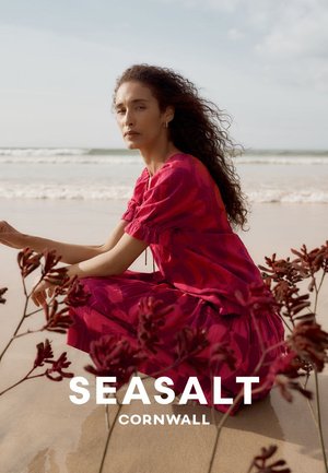 Vrouw met lang krullend haar die een rode jurk draagt, zit op een zandstrand bij zeewier met golven op de achtergrond.