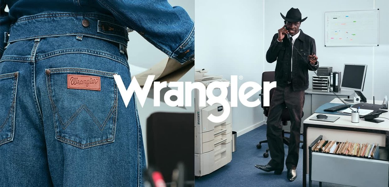 Blaue Jeans aus Denim mit einem Leder-Patch von Wrangler, kombiniert mit einer schwarzen Jacke. Büroumgebung mit einem Drucker, einem Schreibtisch und sichtbaren Unterlagen.