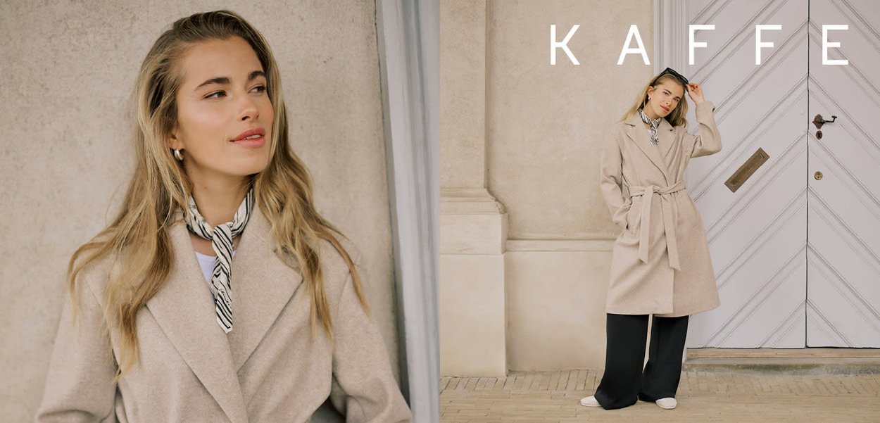 Frau in beigem Trenchcoat, schwarzen weitem Hosen, gestreiftem Halstuch und Sonnenbrille steht bei einer weißen Tür mit dem KAFFE-Logo darüber.