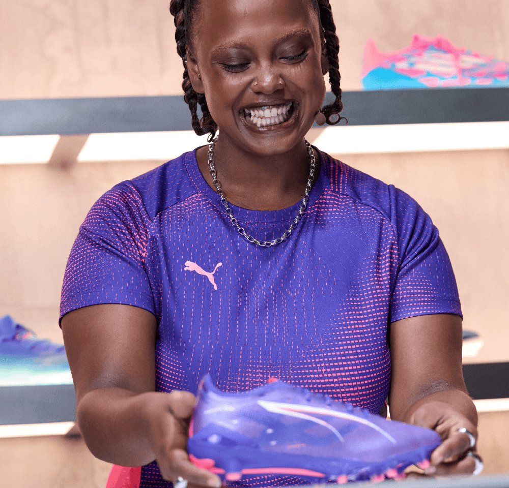 Puma Sportschuhe in Lila mit pinken Akzenten, leichtes Design, glatte Textur und spitze Zehenform, gehalten von einer Person in einem lila Shirt.