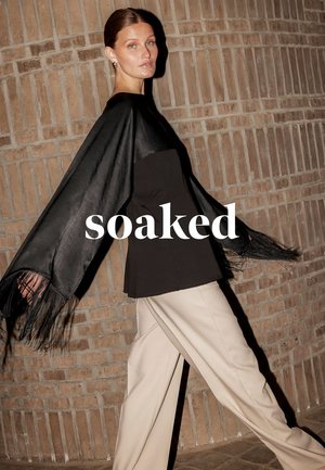 Donna che cammina accanto a un muro di mattoni indossando una giacca nera di camoscio con frange e pantaloni beige, con la scritta "soaked" sovrapposta.