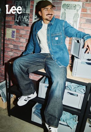 Jeansjacke in hellblau mit Vordertaschen und Lee-Logo; kombiniert mit dunkelblauen Jeans und weißen Sneakern, vor einem Ziegelmauer-Hintergrund.