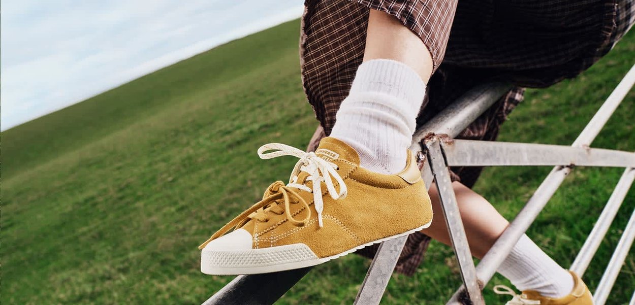 Primer plano de una persona que lleva zapatillas de ante amarillas y calcetines blancos, sentada en una cerca metálica con césped verde al fondo.