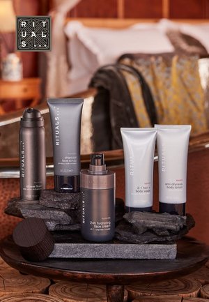 Four Rituals products displayed on dark stone: a gray shower foam, charcoal face scrub, hydrating face cream, and body washes in white tubes.