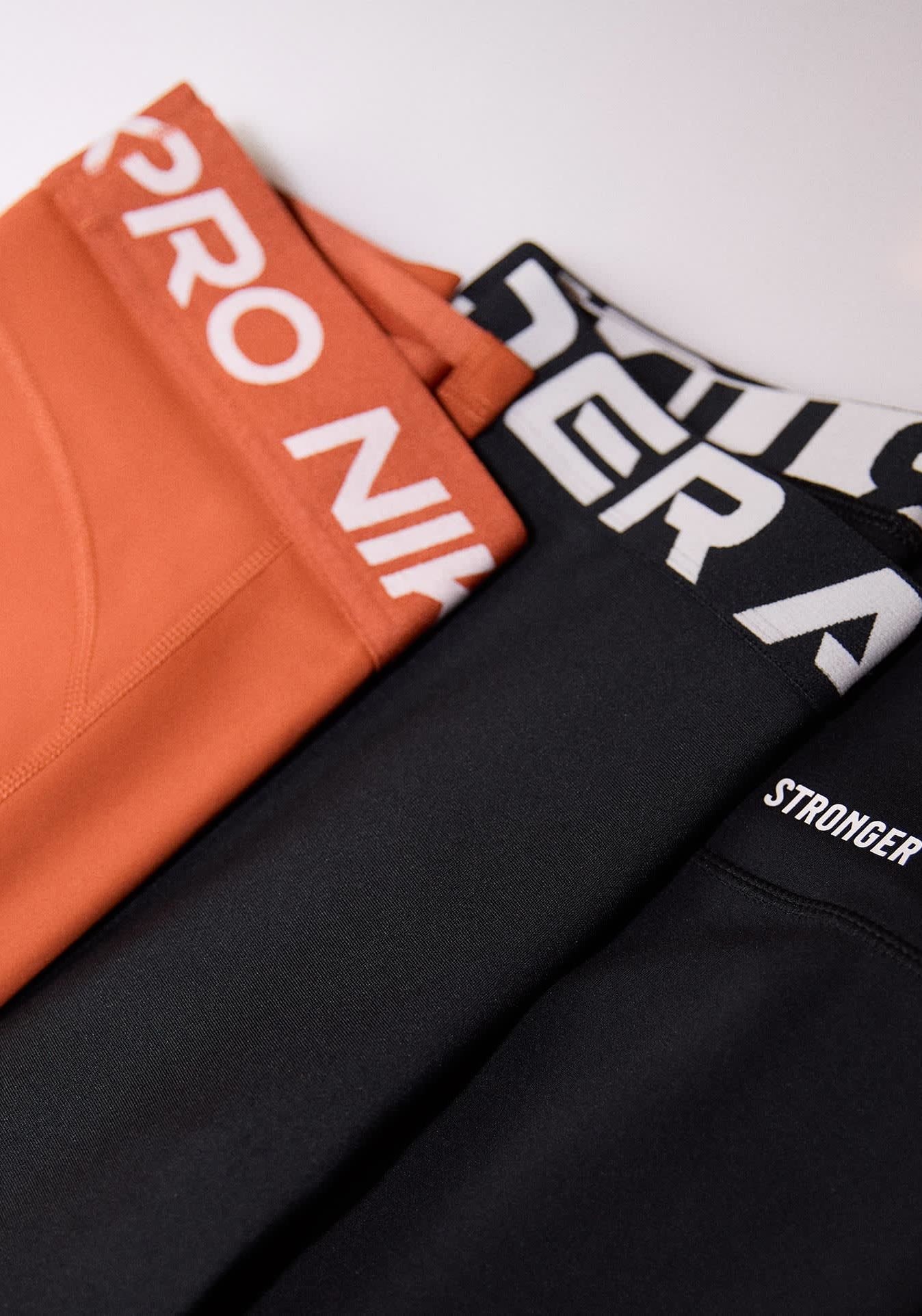 Zwei Paar Leggings: eine orange mit einem weißen "Nike Pro" Bund und eine schwarze mit einem weißen "Stronger" Etikett und gemustertem Bund.