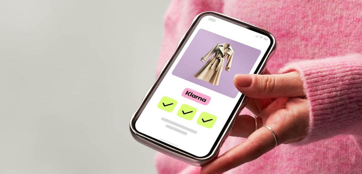 Smartphone, das einen beigen Trenchcoat auf einem pastellfarbenen Hintergrund zeigt. Der Benutzer hält das Telefon in einem sanften rosa Pullover, der Häkchen und "Klarna" zeigt.