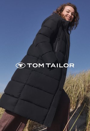 Schwarze Puffermantel mit gestepptem Design, ausgestattet mit Kapuze und frontalem Reißverschluss. Glatte Textur, knielanger Schnitt, getragen über braunen Hosen.