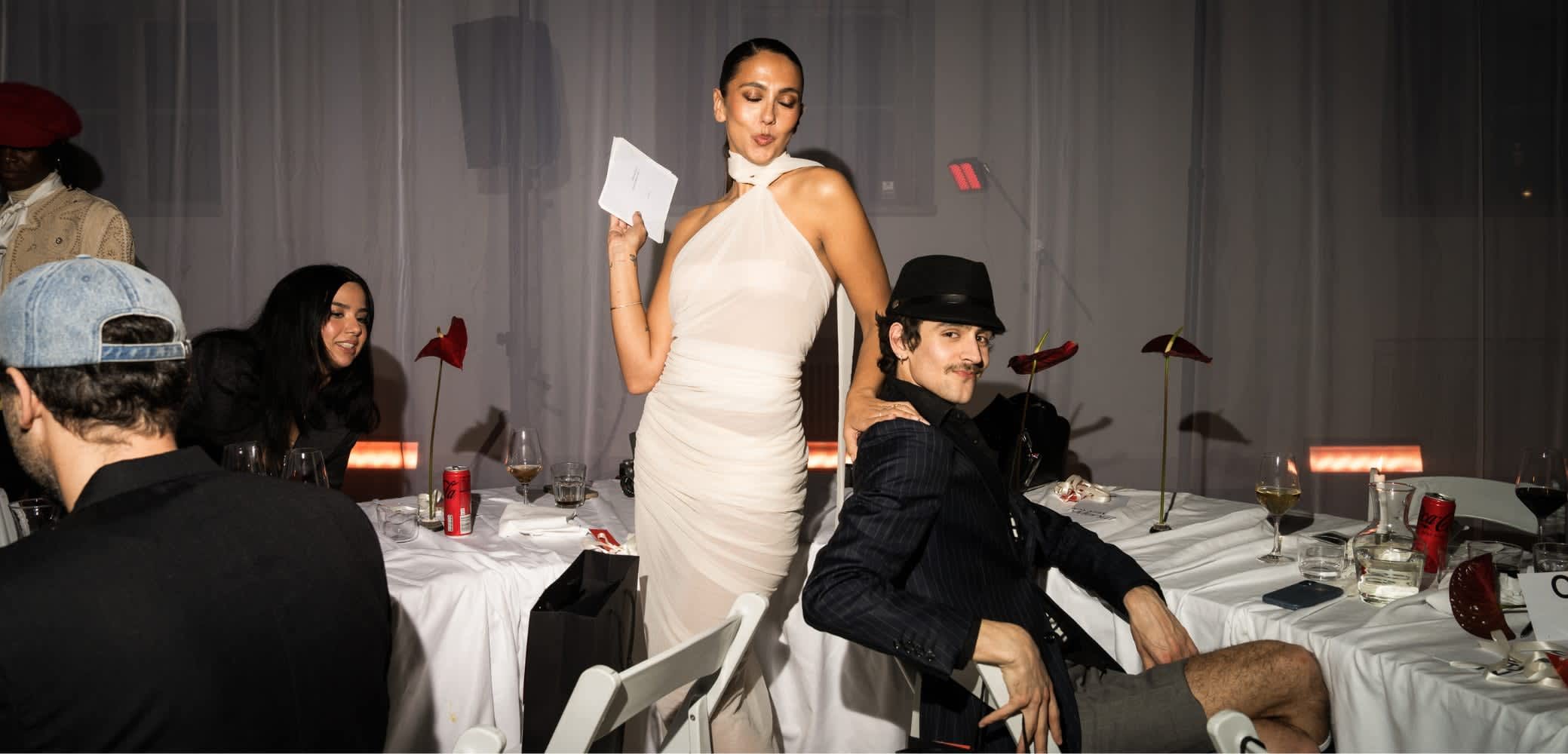 En modell i en draperad beige klänning poserar med ett kort. En man i en svart kavaj med kritstrecksrand och hatt sitter bredvid henne vid ett bord med drycker och dekorationer.