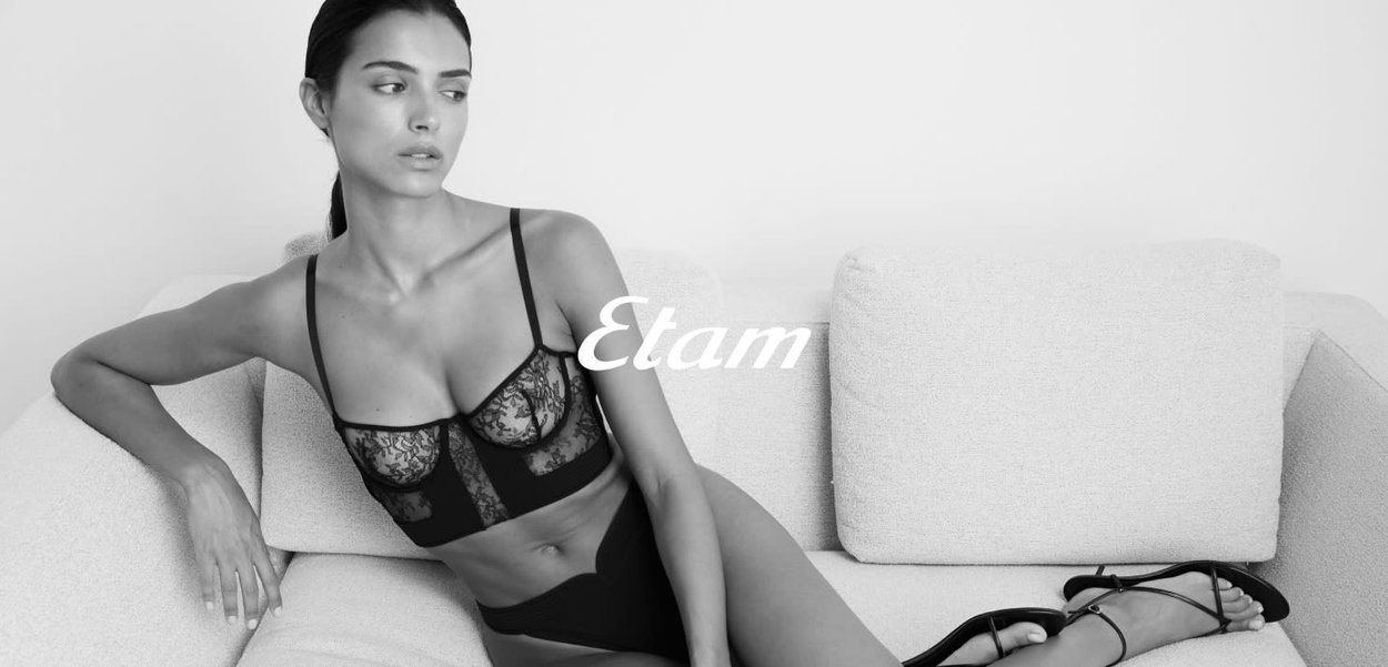 Soutien-gorge en dentelle noire avec des panneaux transparents et des bretelles élastiques, associé à une culotte noire taille haute. Le modèle se détend sur un canapé clair texturé.