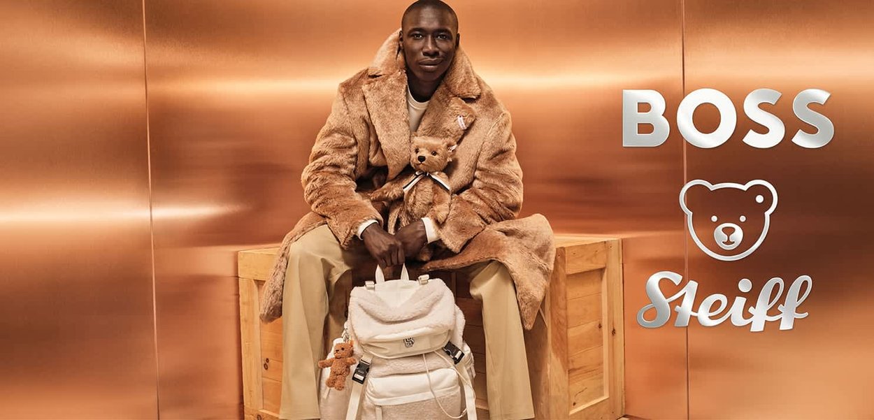 Brauner Kunstfellmantel, cremefarbener Rucksack mit strukturierten Akzenten und ein Teddybär in der Hand, vor einem kupferfarbenen Hintergrund. Logos: BOSS, Steiff.
