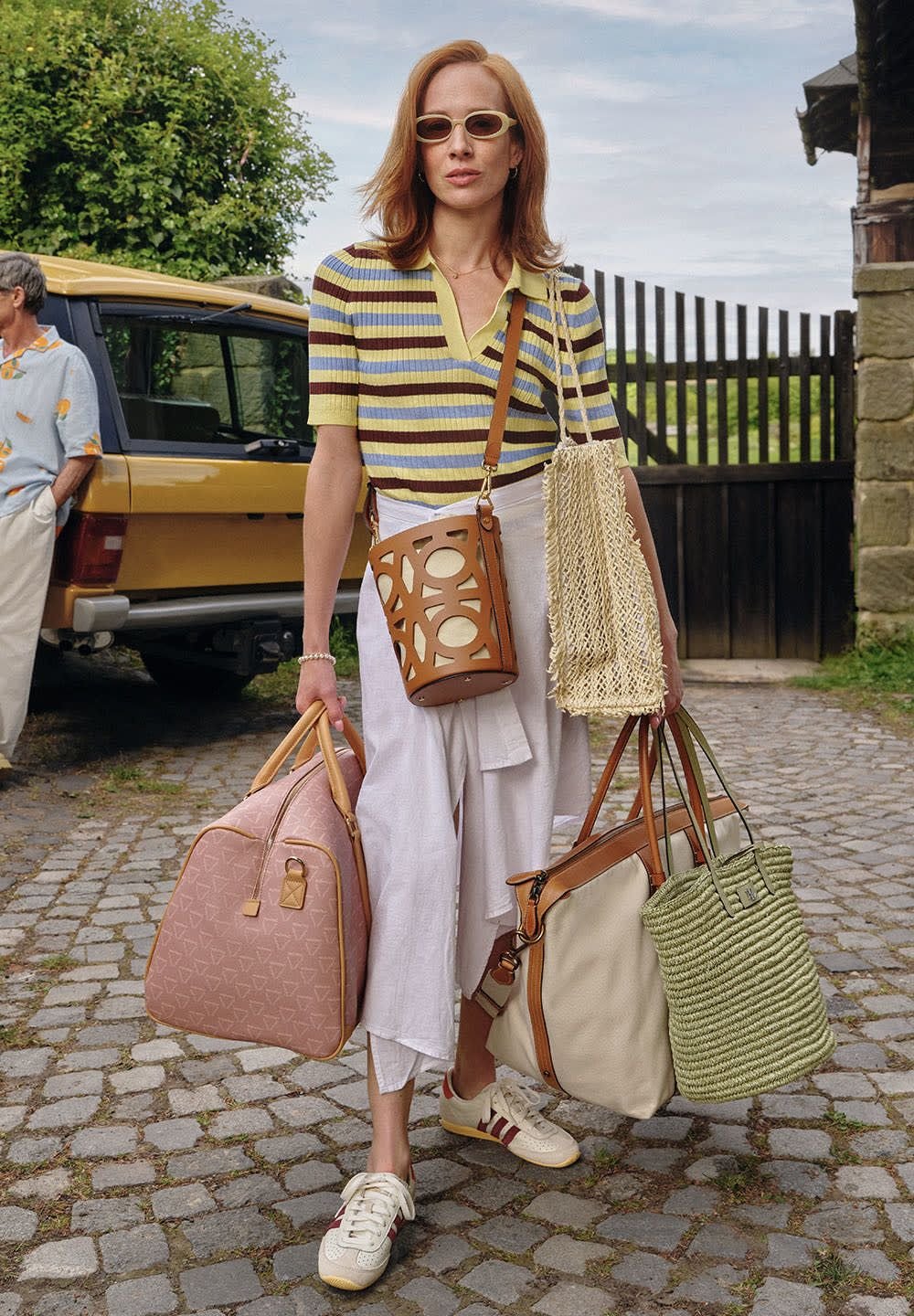 Kabelka s hnědo-tanovým vzorem, růžový cestovní kufr, zelená pletená taška a kulatá hnědá peněženka s vyřezávaným vzorem, všechny neseny. Bíle sukně a pruhovaný top.