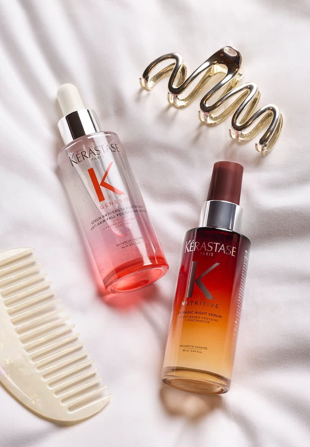Two Kérastase hair serums on white fabric: a clear pink bottle with a dropper and a gradient amber to clear bottle with a pump. A gold hair clip and a white comb are included.
