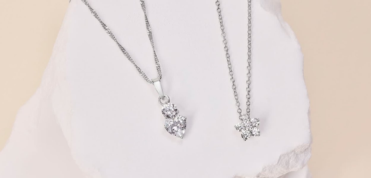 Deux colliers en argent sont exposés sur une surface blanche. L'un présente un pendentif en forme de cœur avec trois pierres précieuses transparentes, l'autre un pendentif en forme d'étoile avec quatre pierres transparentes.