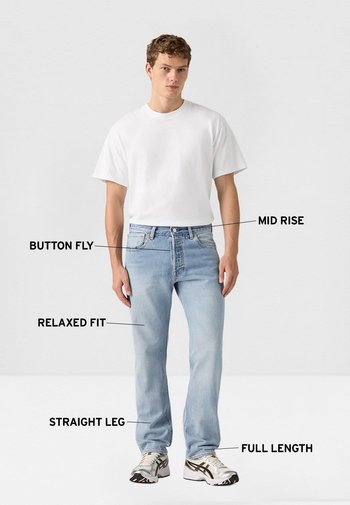 Joven con jeans azules claros de corte recto y talle medio con cierre de botones y camiseta blanca, de pie contra un fondo liso.