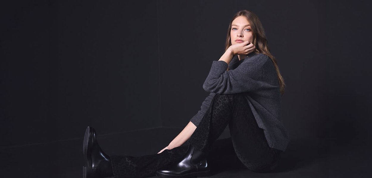 Bottines Chelsea noires, pantalon en velours texturé foncé à imprimé léopard, et un pull tricoté gris oversized. Mannequin assis sur un fond sombre.