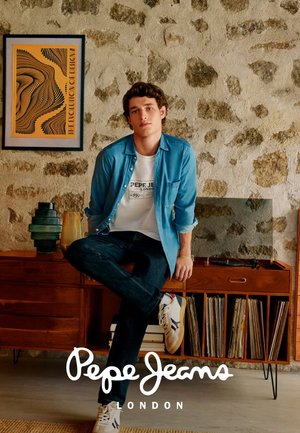 Jongeman in een casual denim overhemd, wit graphic T-shirt en sneakers zit op een mid-century houten kast met platen en decoratieve vaasjes.