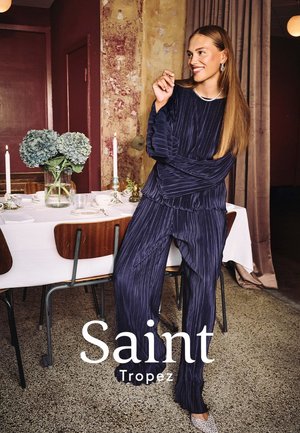 Navy pleated outfit featuring long sleeves and relaxed pants, paired with textured, light-colored shoes. Green floral arrangement on table.