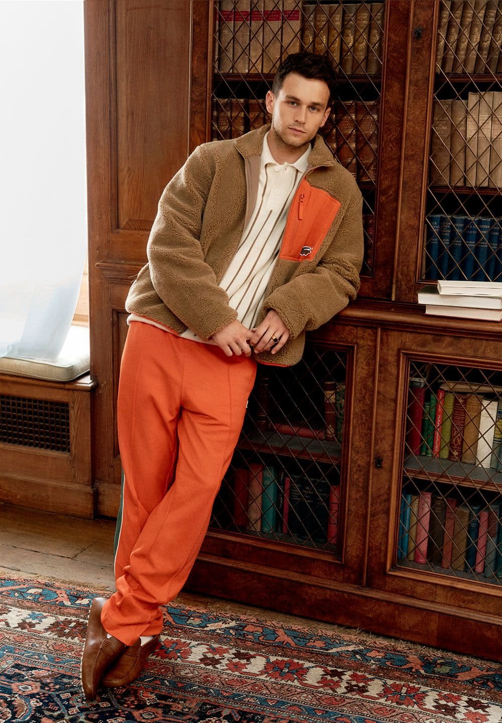 Veste en polaire marron avec une poche orange, polo à rayures blanches et pantalon de survêtement orange. Chaussures marron. Bibliothèque en bois en arrière-plan.