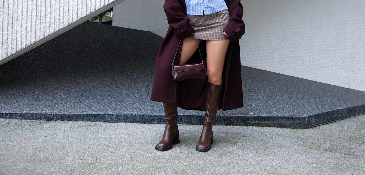Stivali alti fino al ginocchio in pelle marrone con tacco a blocco, abbinati a una minigonna scozzese e a un lungo cappotto color bordeaux, su uno sfondo grigio texture.