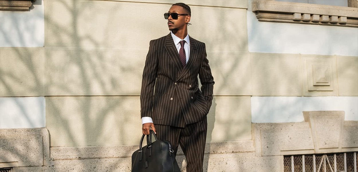 Hombre con traje de rayas oscuras y gafas de sol, sosteniendo un maletín de cuero negro, de pie frente a una pared de piedra iluminada por el sol al aire libre.