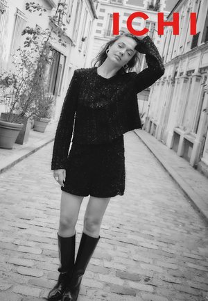 Schwarzes, strukturiertes zweiteiliges Outfit bestehend aus einem kurzen Pullover und Shorts. Das Modell trägt kniehohe schwarze Stiefel und silberne Creolen.