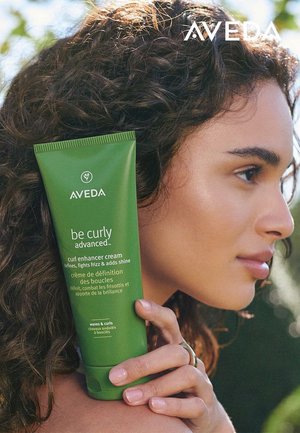 Grønn tube med Aveda curl enhancer cream holdt ved siden av ansiktet til en person. Teksten inkluderer "be curly advanced" og produktfordeler.