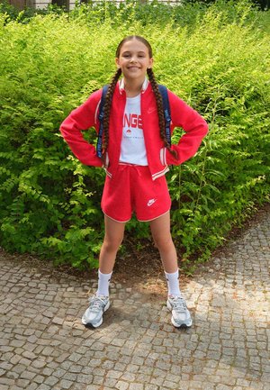 Ragazza giovane con capelli intrecciati indossa giacca sportiva rossa e pantaloncini, maglietta bianca e scarpe da ginnastica, in piedi con sicurezza su un sentiero di ciottoli con cespugli verdi dietro.