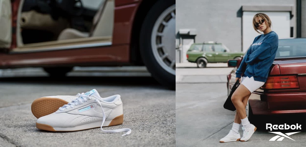 Zapatillas blancas Reebok sobre el pavimento junto a un coche rojo; una persona con zapatillas, calcetines blancos, pantalones cortos y sudadera azul se apoya en el coche.
