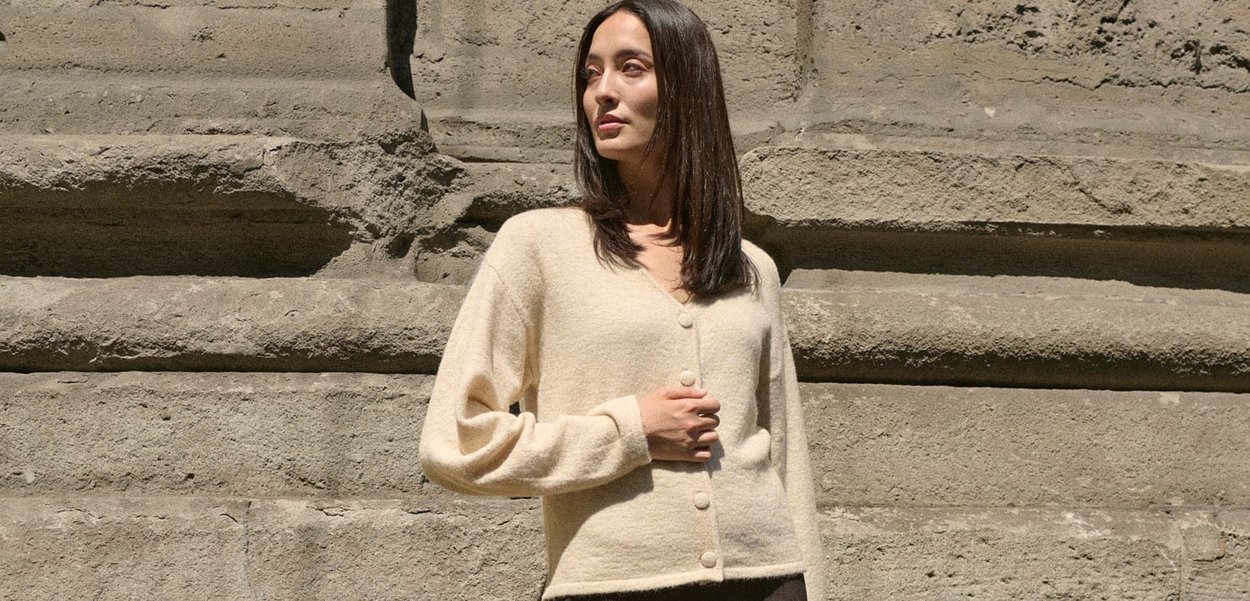 Cardigan beige realizzato in materiale morbido, caratterizzato da un collo a V, maniche lunghe e cinque bottoni sul davanti. Texture liscia con dettagli minimi.